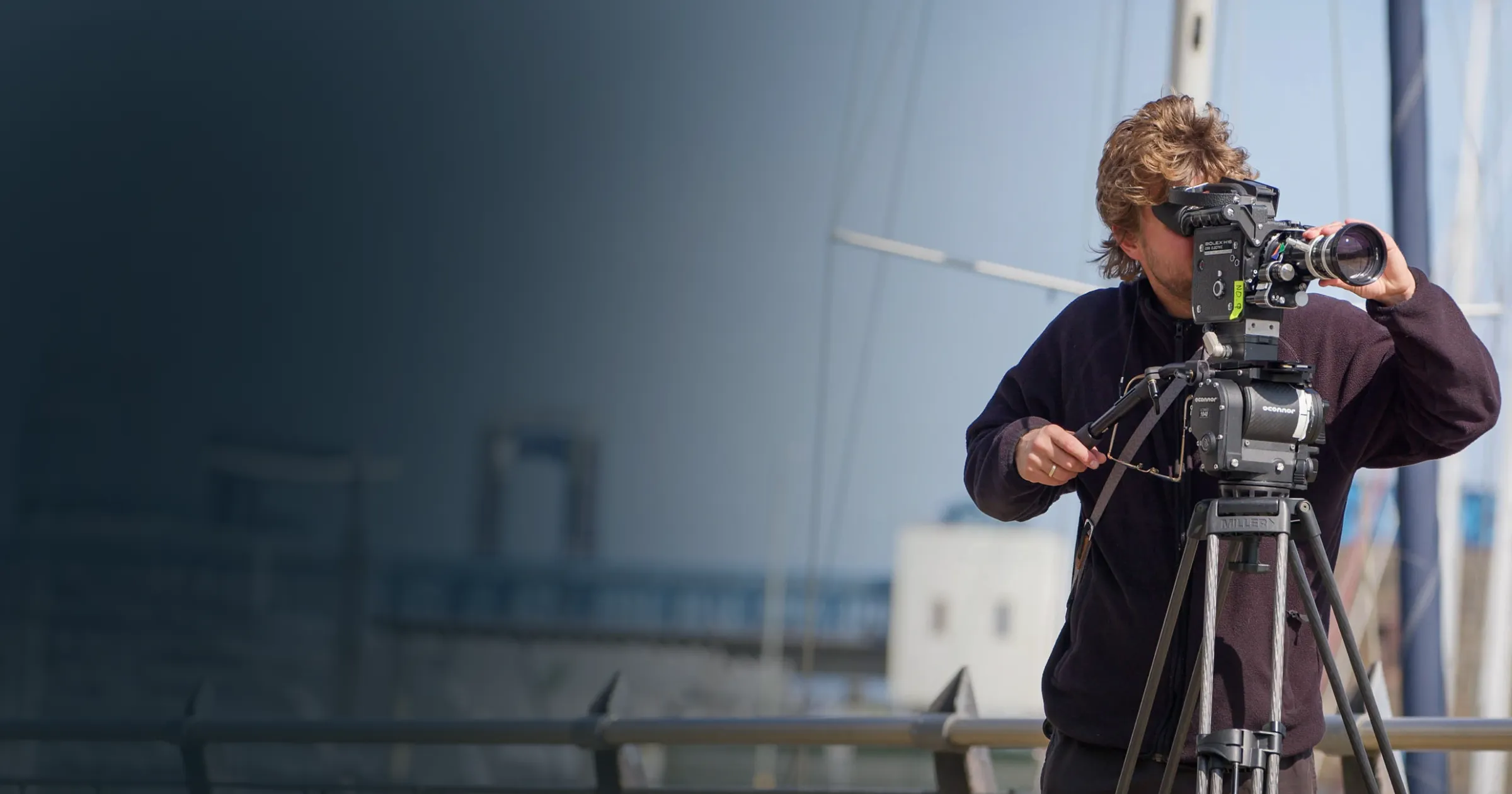 A person operating a camera on a tripod outdoors, focusing intently. The background features a blurred harbor scene, creating a professional, focused mood.